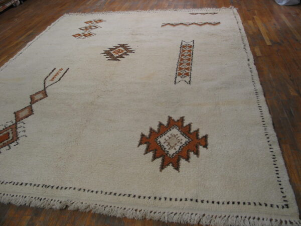 Vintage room size rabat tribal moroccan berber ivory wool color rug 4 Off-white shaggy rug with orange and brown geometric motifs placed on a dark wood floor.
