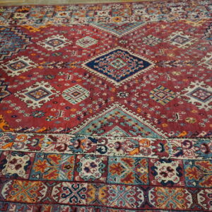 Angled view of a vibrant red rug with a central diamond medallion and multi-colored geometric borders.