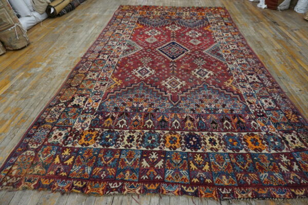 Deep red rug with complex blue and gold geometric designs lies on light distressed wood flooring.