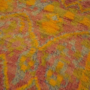 High-pile rug with geometric shapes in orange-yellow, reddish-pink, and pale blue on a wood floor.
