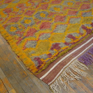 Multicolored high-pile rug with geometric shapes, flat-weave border, and purple fringe on wood floor.