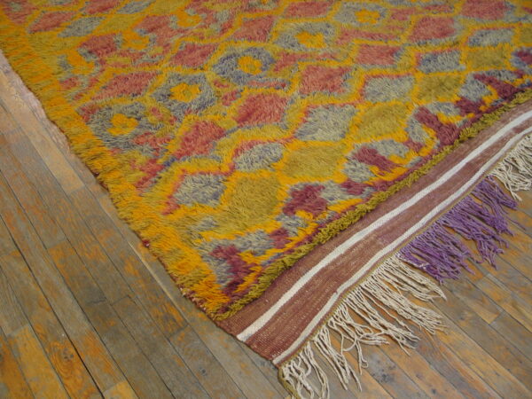 Multicolored high-pile rug with geometric shapes, flat-weave border, and purple fringe on wood floor.