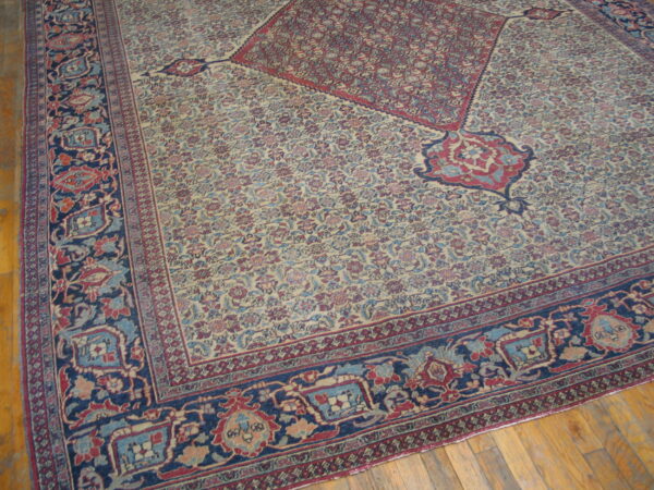 Intricate low-pile rug featuring red, blue, and cream floral designs, a navy border, and corner motifs on wood flooring.
