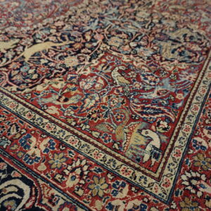 Intricate low-pile rug corner featuring red, navy, and cream colors with detailed bird and animal patterns.