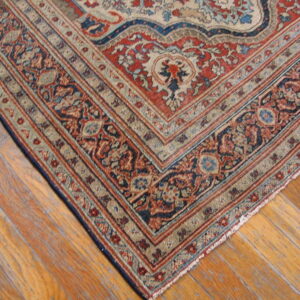 Close-up of an ornate rug corner in red, navy blue, and cream borders resting on wood plank flooring.