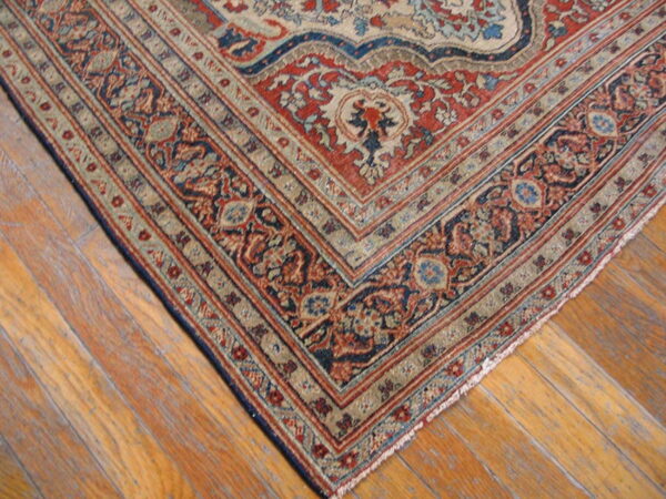 Close-up of an ornate rug corner in red, navy blue, and cream borders resting on wood plank flooring.