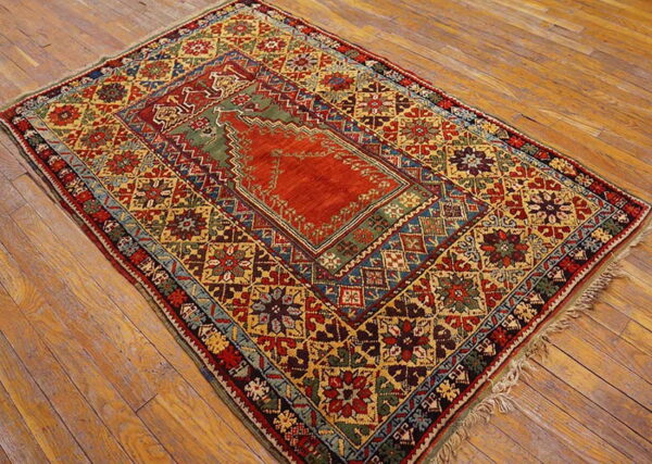 Low-pile rug with rust and olive geometric patterns and intricate borders rests on light brown wood flooring.