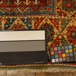 High-pile rug texture with complex red, gold, teal, and dark green patterns; visible fringe and wood floor.