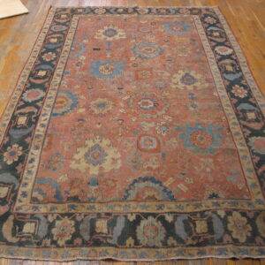 Muted red-orange rug with an intricate blue and tan floral pattern and a dark border on wood flooring.