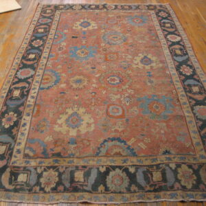 Rectangular rug featuring a muted coral field, complex floral designs, and a wide dark border on wood plank flooring.
