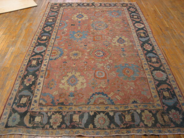 Rectangular rug featuring a muted coral field, complex floral designs, and a wide dark border on wood plank flooring.