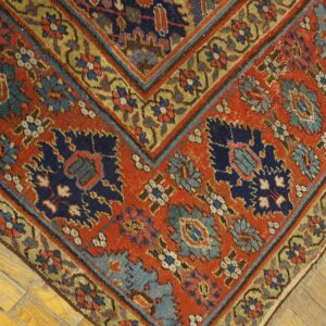 Ornate rug corner showing rust red, navy, and gold geometric motifs over distressed yellow wood flooring.