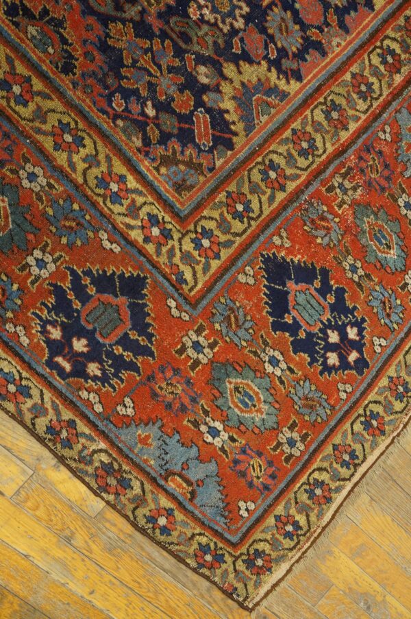 Ornate rug corner showing rust red, navy, and gold geometric motifs over distressed yellow wood flooring.