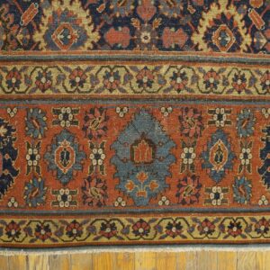 Rust and navy traditional rug showing intricate geometric borders on light wood floorboards.