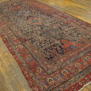 Ornate traditional low-pile rug featuring a red border and dark blue field on worn wooden planks.