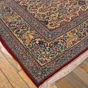 Beige rug corner with dense blue, red, and tan traditional pattern and a dark red edge on wood planks.