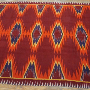 Deep red geometric rug featuring repeating serrated orange and blue diamond patterns on a wood floor.