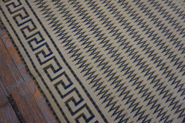 Flat-weave rug featuring a beige field, blue and brown zigzag pattern, and geometric border on wood planks.