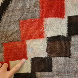 A finger highlights a hole in a flat-woven geometric rug patterned with red, cream, dark brown, and grey blocks.