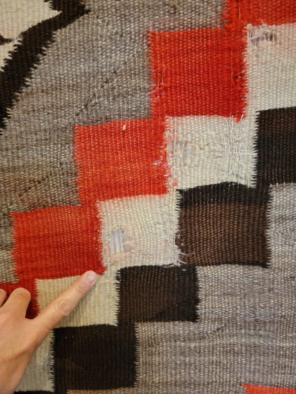 A finger highlights a hole in a flat-woven geometric rug patterned with red, cream, dark brown, and grey blocks.