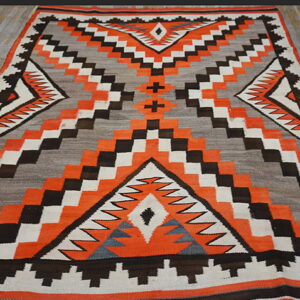 Geometric flatweave rug featuring orange, black, white, and gray patterns, displayed on a plank wood floor.