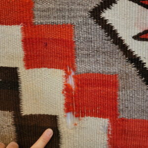 Detailed flatweave rug showing orange, cream, brown, and gray stepped patterns with damage; a finger points below.
