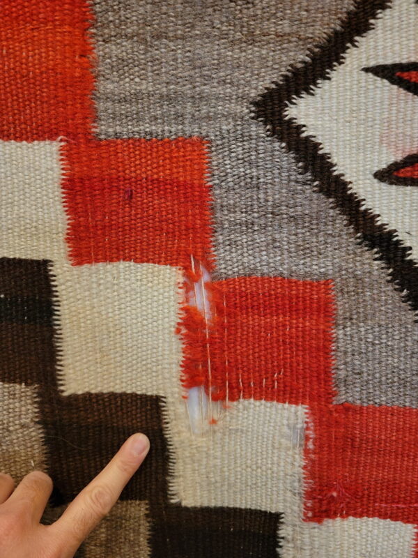 Detailed flatweave rug showing orange, cream, brown, and gray stepped patterns with damage; a finger points below.