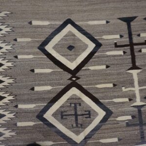 Gray flatweave rug with dark geometric diamonds, crosses, and arrow motifs on wood flooring.