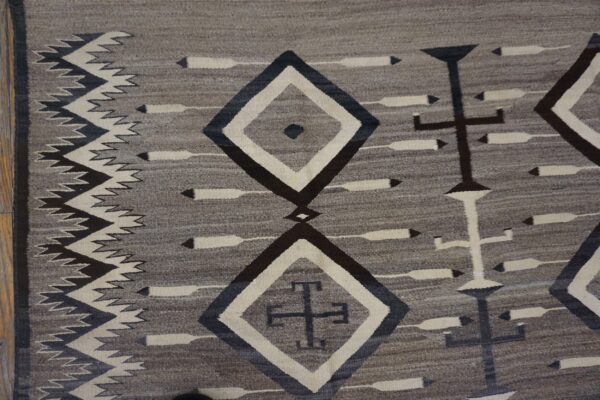 Gray flatweave rug with dark geometric diamonds, crosses, and arrow motifs on wood flooring.