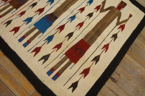 Cream and brown flatweave rug with stylized human figures, red and blue arrows, and a black border on wood planks.