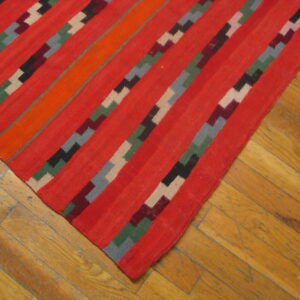 Flatweave rug with diagonal red, orange, and stepped geometric stripes on worn wood floors.