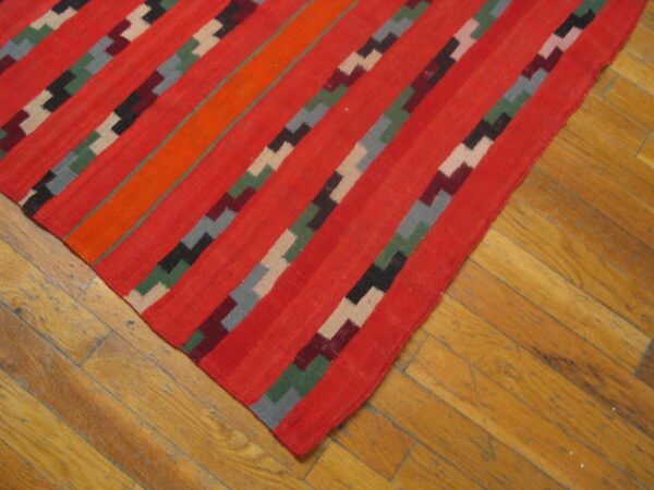 Flatweave rug with diagonal red, orange, and stepped geometric stripes on worn wood floors.