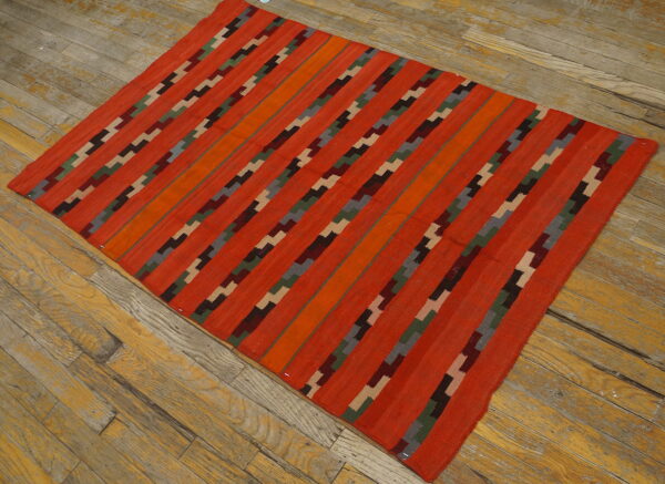 Red flatweave rug with dark, multi-colored stepped stripe patterns lying diagonally on distressed wood floor.