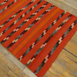 Bright orange and red flatweave rug with geometric stepped diagonal stripes rests on distressed wood flooring.