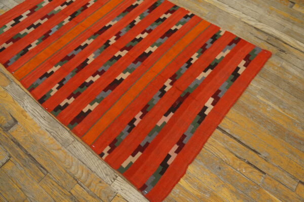 Bright orange and red flatweave rug with geometric stepped diagonal stripes rests on distressed wood flooring.