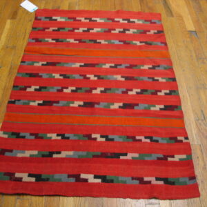 Red flatweave rug with horizontal stripes and multicolored stepped geometric patterns on hardwood flooring.