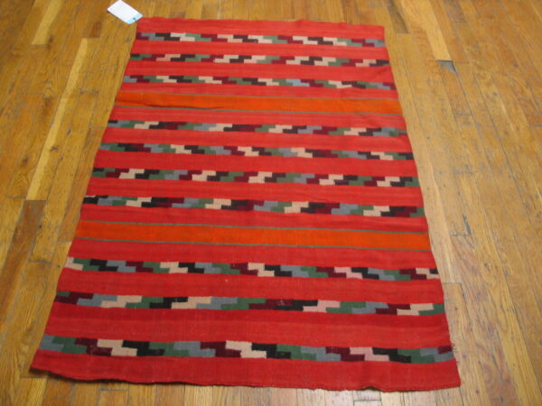 Red flatweave rug with horizontal stripes and multicolored stepped geometric patterns on hardwood flooring.