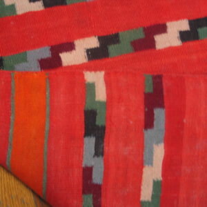 Flatwoven rug featuring red fields and multicolored geometric bands, folded over light wood flooring.
