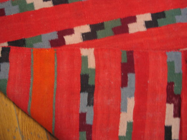 Flatwoven rug featuring red fields and multicolored geometric bands, folded over light wood flooring.