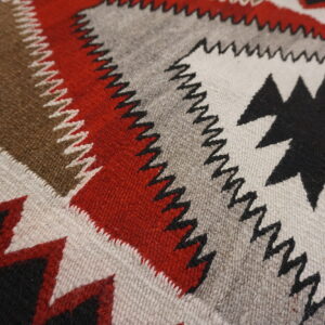 Close-up of a flatweave rug with bold geometric bands and serrated edges in red, black, gray, and brown.