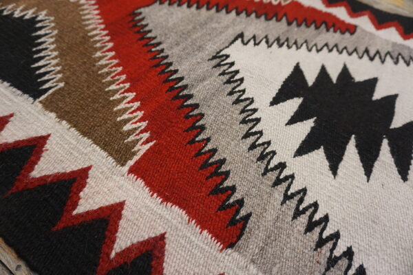Close-up of a flatweave rug with bold geometric bands and serrated edges in red, black, gray, and brown.