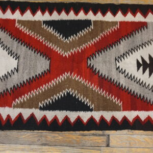 Flatwoven rug in red, black, white, gray, and brown geometric zigzag patterns on a rustic wooden floor.