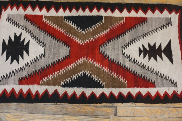 Flatwoven rug in red, black, white, gray, and brown geometric zigzag patterns on a rustic wooden floor.