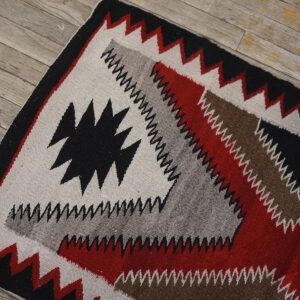Geometric flat weave rug with black, red, gray, and brown zigzag patterns rests on weathered wood planks.