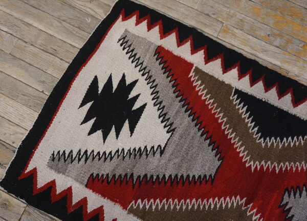 Geometric flat weave rug with black, red, gray, and brown zigzag patterns rests on weathered wood planks.