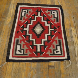 Red, black, white, and gray flat-weave geometric rug placed on worn plank hardwood flooring.