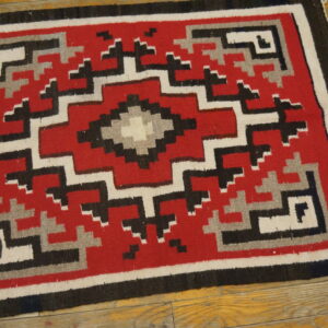 A flat-weave rug with a red, black, gray, and white geometric diamond pattern on a wooden floor.