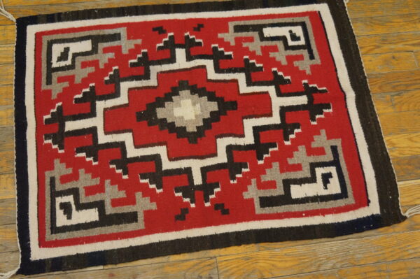 A flat-weave rug with a red, black, gray, and white geometric diamond pattern on a wooden floor.