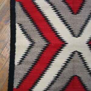 Close-up of a low-pile rug with red, gray, and cream serrated geometric designs on wood flooring.