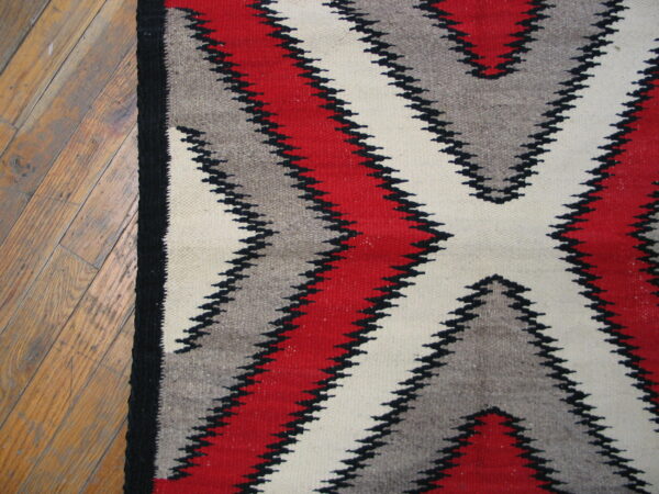Close-up of a low-pile rug with red, gray, and cream serrated geometric designs on wood flooring.